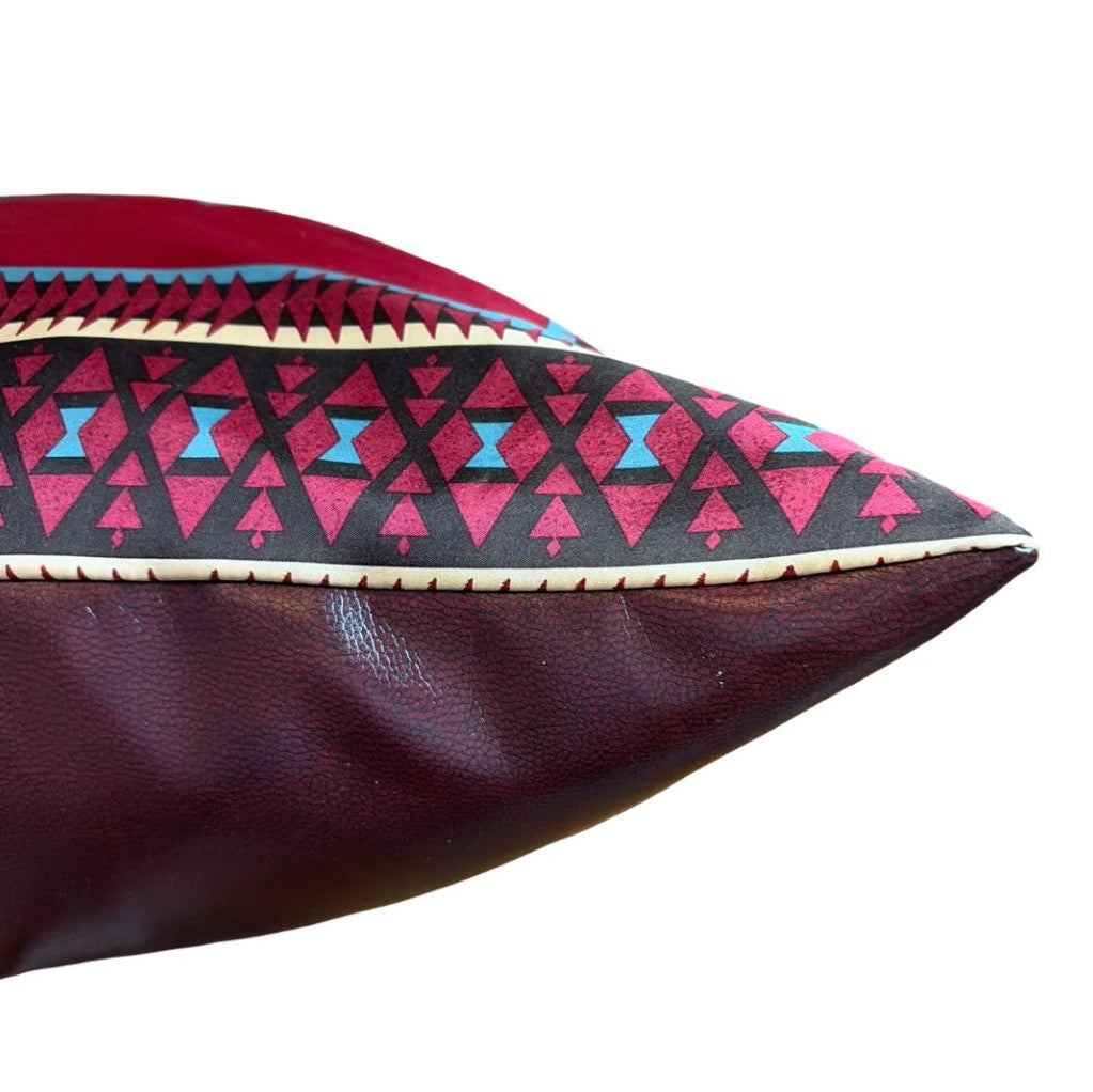A close-up of a decorative throw pillow featuring a dark burgundy leather-like base, a black geometric patterned section with pink and light blue hourglass triangles, and a cream stitched border.