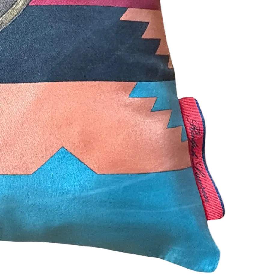 A close-up of a colorful scarf with a geometric pattern in navy blue, peach, and turquoise, featuring a pink woven brand tag on the edge.