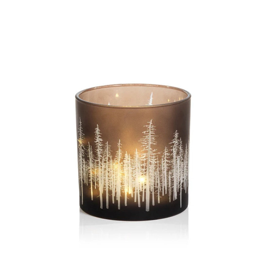 A lit candle in a dark brown glass holder featuring a white etched forest pattern, shown against a plain white background.