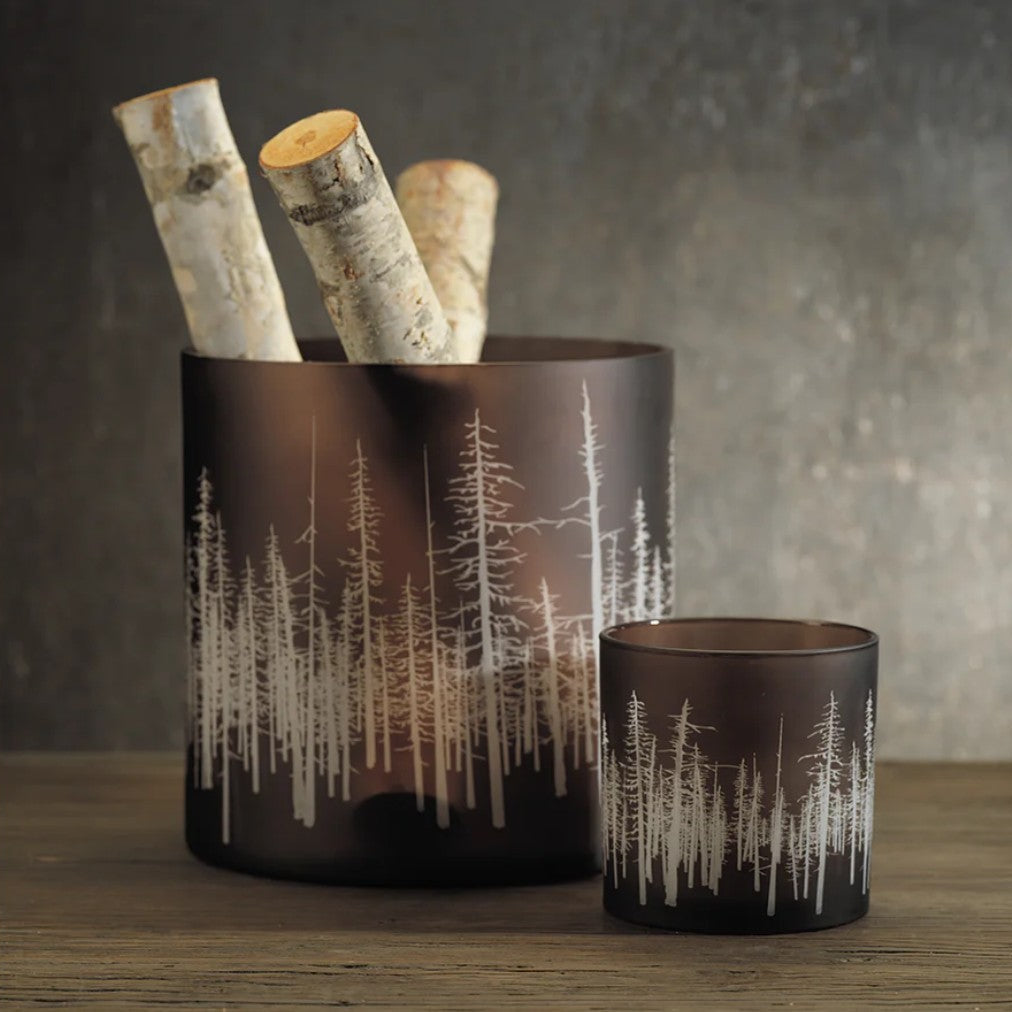 Two frosted dark brown glass candle holders with white etched pine tree silhouettes, one larger container holding birch logs and one smaller matching candle holder, set on a wooden surface against a textured gray background.
