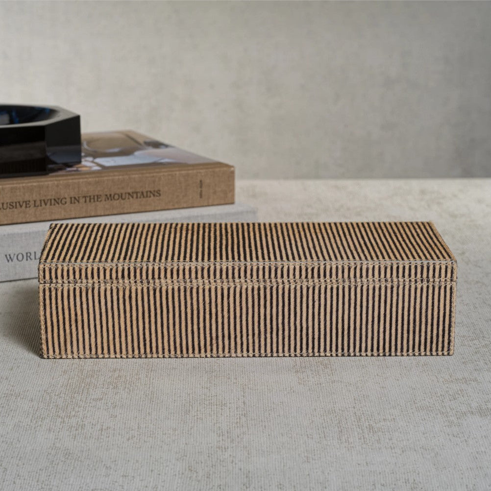 A rectangular wooden box with a slatted, ribbed texture in a light brown wood tone, displayed on a textured beige fabric surface with a neutral gray background and decorative books behind it.
