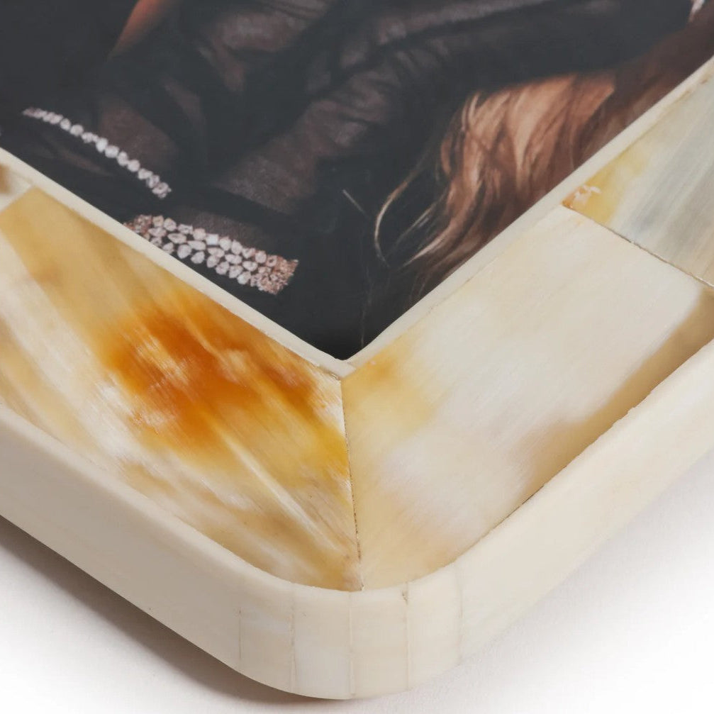 Close-up corner view of a rectangular tray or small table with a printed artwork on top. The base has a cream and amber marbled pattern, and the top surface shows a dark fashion photograph with a jeweled bracelet and hair.
