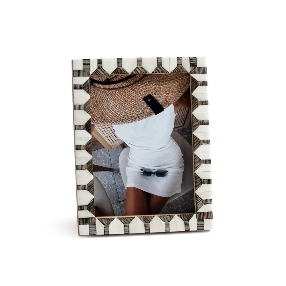 A rectangular photo frame with a geometric black and white patterned border, displaying a photo of a woman in a white dress and straw hat, set against a plain white background.