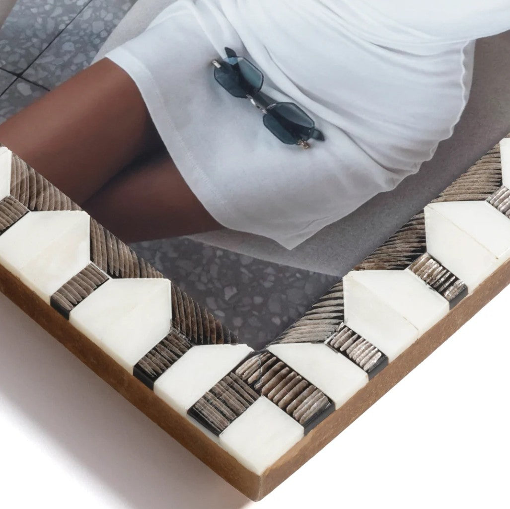 A close-up of a geometric framed wall art piece with white and gray stone-like shapes and woven brown strips, shown in a styled interior setting with part of a person's legs and white skirt visible.