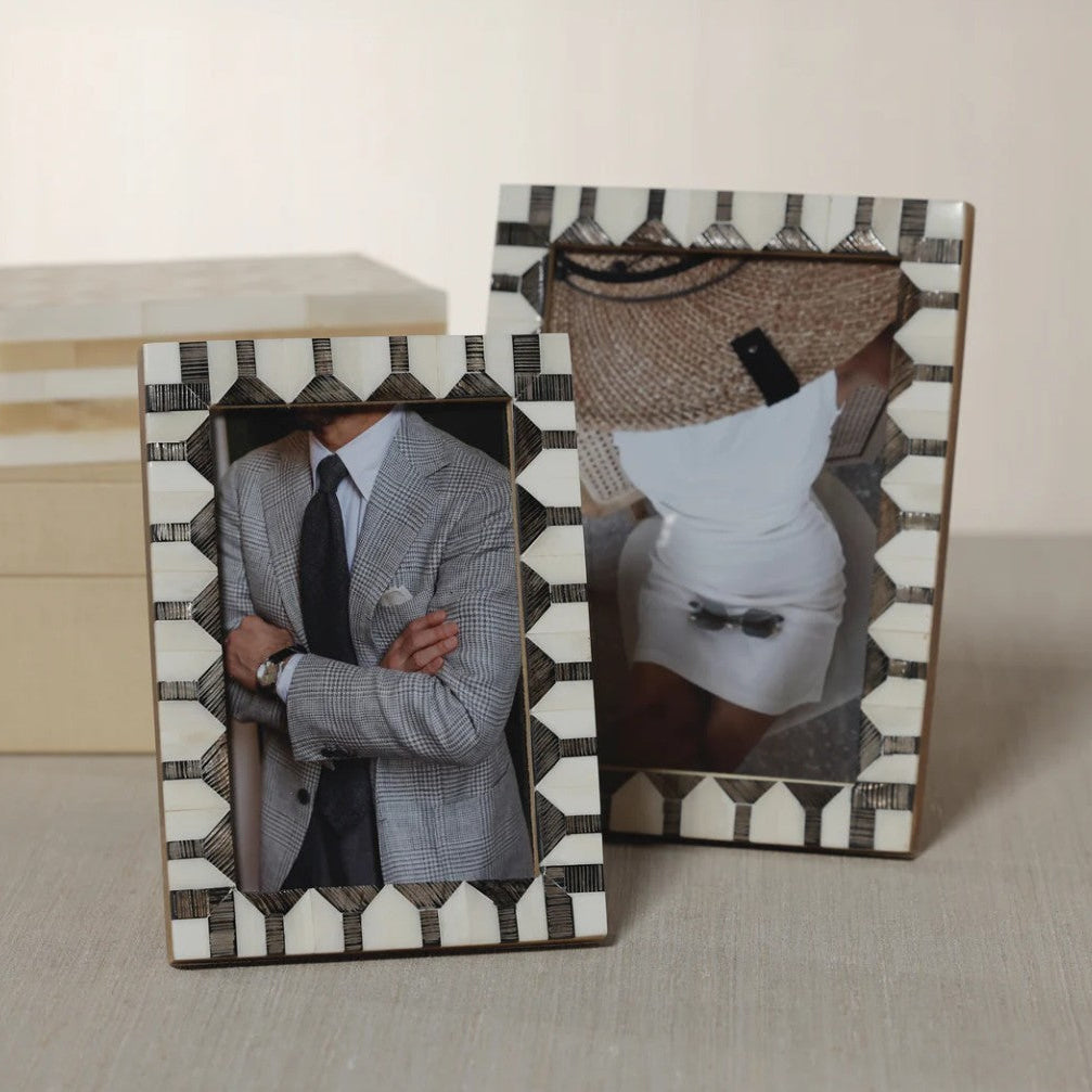 Two rectangular picture frames with a black-and-white geometric shell-like border, displayed on a light beige surface. The left frame contains a photo of a man in a gray suit, and the right frame contains a photo of a person wearing a white dress and a woven hat.