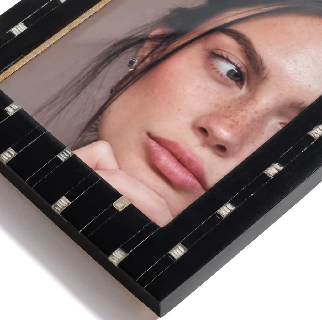 A close-up of a black picture frame with a glossy finish, holding a portrait of a woman with freckles. The frame has small decorative silver-toned accents along the inner border.