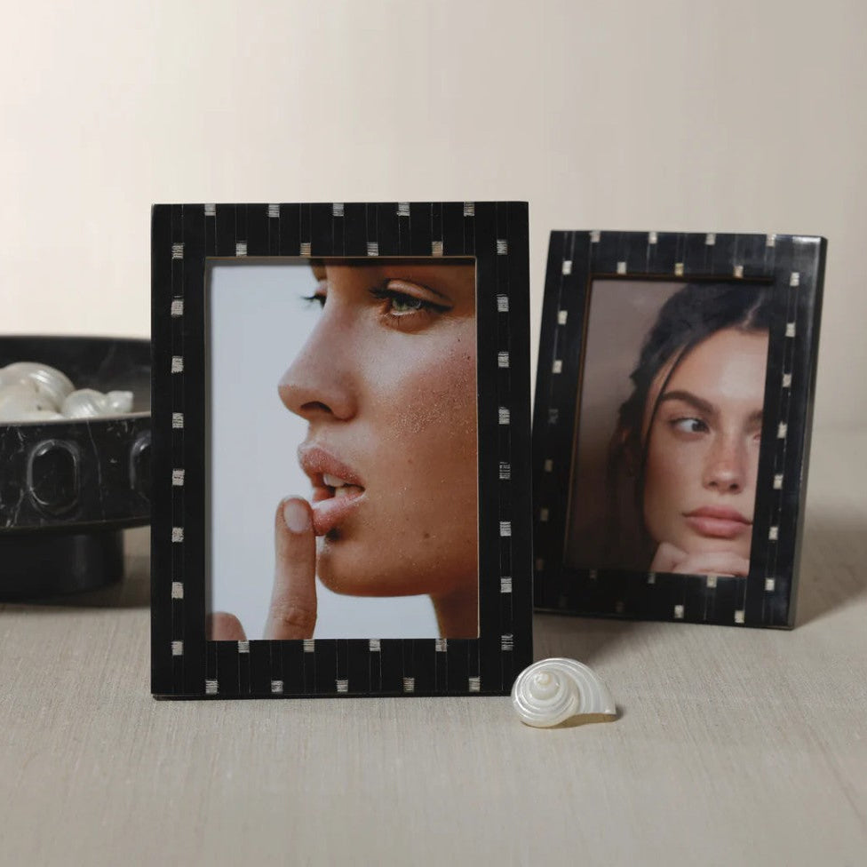 Two black picture frames with a small white geometric pattern, displayed on a light beige surface. The larger frame in the foreground displays a close-up portrait of a woman, while the smaller frame behind it displays another portrait.